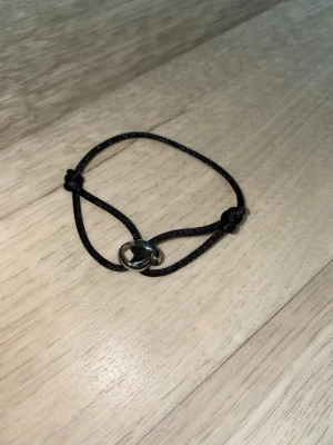 Svart armband med silverfärgad ring - Snyggt armband med svart justerbart band och en stilren silverfärgad ring i mitten. Perfekt accessoar för en enkel men trendig look. Passar att bära ensam eller tillsammans med andra armband för en personlig stil.