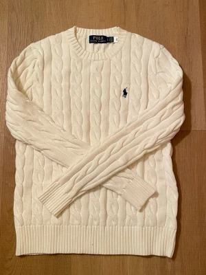 Vit kabelstickad tröja Polo Ralph Lauren - Kabelstickad vit tröja från Polo Ralph Lauren med klassisk rund halsringning och broderad marinblå logga på bröstet. Tröjan har ribbade muddar och nederkant, perfekt för en stilren look. Materialet är mjukt och bekvämt.