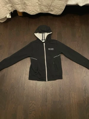 Svart hoodie från Hugo Boss - Snygg svart hoodie från Hugo Boss med vit dragkedja och logga på bröstet. Hoodien har huva med ljusgrå insida och två fickor framtill. Perfekt för en chill och stilren look. Materialet känns mjukt och bekvämt.