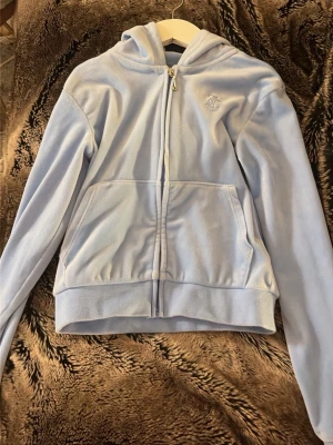 Ljusblå velour hoodie från Juicy Couture - Säljer en ljusblå hoodie i mjuk velour från Juicy Couture. Hoodien har dragkedja framtill, två stora fickor och en broderad logga på bröstet. Klassisk huva och ribbade muddar vid ärmar och nederkant. Perfekt för en chill och trendig look.