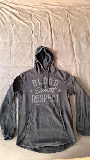 Mörkblå hoodie från Under Armour - Snygg mörkblå hoodie från Under Armour med trycket 'Blood Sweat Respect' på bröstet. Hoodien har huva med dragsko och en stor magficka. Tillverkad i mjukt och bekvämt material, perfekt för chill eller träning. Loose fit för extra komfort.