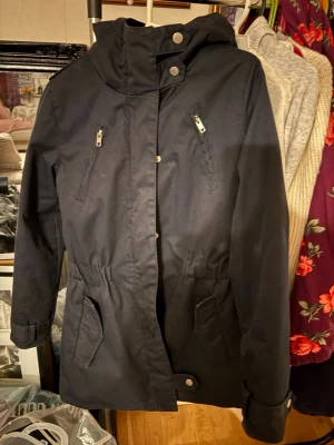 Marinblå parkas från Elvine med huva - Marinblå parkas från Elvine med quiltat foder, dragkedja och knappar framtill. Jackan har två bröstfickor med dragkedja, två stora fickor nertill och justerbar midja. Perfekt för kyliga dagar med sin stilrena look och praktiska huva.