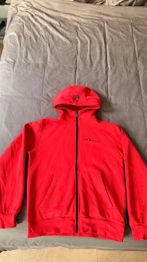 Röd hoodie från Sail Racing - Riktigt snygg röd hoodie från Sail Racing med dragkedja, två stora fickor fram och en unik logga på huvan. Tillverkad i mjukt material. Perfekt för dig som vill ha en stilren och sportig look. Använd 3-4ggr. 