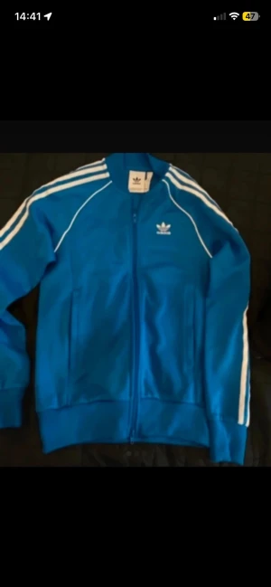 Blå Adidas track jacket - Säljer en klassisk blå Adidas track jacket med vita ränder längs ärmarna och dragkedja framtill. Jackan har ståkrage, två fickor och Adidas logga på bröstet. Perfekt för sport eller chill. Materialet är troligtvis polyester.