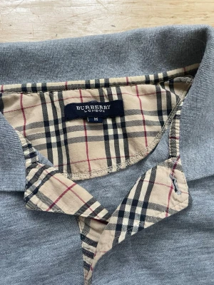 Grå Burberry skjorta med rutig krage - Säljer en grå skjorta från Burberry med klassisk beige, svart och röd rutig krage och knappslå. Skjortan har korta ärmar och en liten röd logga broderad på bröstet. Perfekt för dig som gillar stilrena detaljer och ikoniskt mönster.