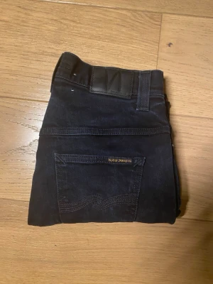 Svarta jeans från Nudie Jeans - Säljer ett par svarta jeans från Nudie Jeans med klassisk femficksmodell och diskret logga på bakfickan. Jeansen har raka ben och är tillverkade i ett slitstarkt denimtyg. Perfekta för en clean och stilren look.