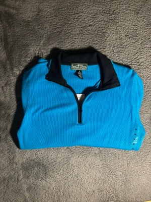 Blå halvzip från Lauren Ralph Lauren - Snygg blå halvzip-tröja från Lauren Ralph Lauren med svart krage och dragkedja framtill. Tillverkad i bomull och har en sportig vibe som passar perfekt till jeans eller träningsbyxor. Enkel men stilren design med ribbad struktur.