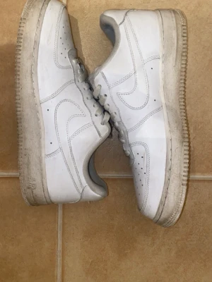Nike Air Force 1 vita sneakers med crease skydd - Klassiska vita Nike Air Force 1 sneakers i lågt utförande. Skorna har en ikonisk siluett med perforerad tå, vit sula och tydlig Swoosh-logga på sidan. Tillverkade i läder med snörning och vadderad kant för extra komfort.