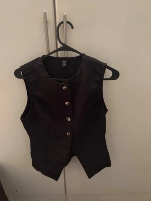 Svart väst med knappar - Stilren svart väst med fyra silvriga knappar framtill och rund halsringning. Västen har en klassisk passform och är ärmlös, perfekt att styla över en skjorta eller t-shirt. Materialet ser ut att vara bomull eller en bomullsblandning. För mer bilder/justering av pris skriv gärna❤️🤗
