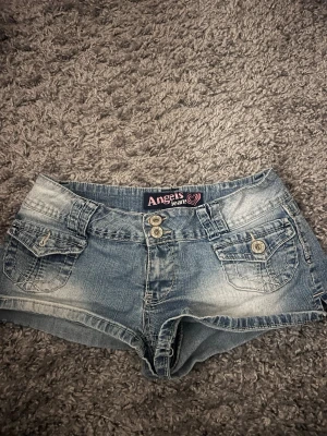 Snygga lågmidjade jeansshorts ❤️‍🔥 - Snygga blå jeansshorts från Angels Jeans med dubbla knappar framtill och fyra fickor. Shortsen har en klassisk denimlook med slitningar och kontrastsömmar. Perfekta för varma dagar och enkel att matcha med olika toppar.