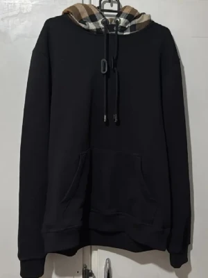 Svart hoodie med rutmönstrad huva - Svart hoodie med klassisk känguruficka och en huva fodrad i beige, svart och röd rutmönstrad textil. Justerbara snören i huvan och ribbade muddar vid ärmslut och nederkant. Perfekt för en chill och stilren look. Storlek S men sitter som xs. Kvitto finns från Farfetch!