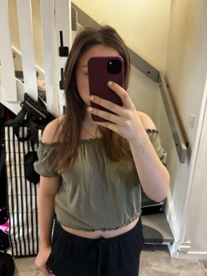 Grön offshoulder croppad topp - Säljer en militärgrön croppad topp med offshoulder-modell och korta ärmar. Toppen har resår och volangdetaljer vid halsringning och nederkant för en söt och avslappnad look. Perfekt till sommaren och enkel att matcha med jeans eller kjol.💖