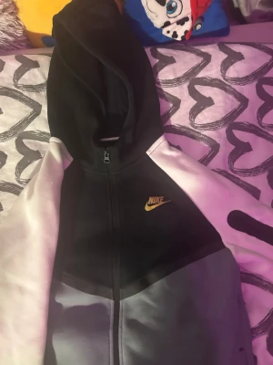 Nike tech hoodie, helt ny typ har använt den 2 gånger bara 158-170cm - Snygg hoodie från Nike med dragkedja framtill. Färgblockad design i svart, grått och ljusrosa med gul Nike-logga på bröstet. Huva och långa ärmar med svart detalj på ena ärmen. Perfekt för en chill och sportig look.