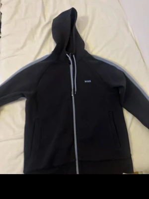 Svart hoodie med dragkedja från BOSS - Snygg svart hoodie från BOSS med dragkedja framtill och huva. Diskret BOSS-logga på bröstet och gråa detaljer längs ärmarna. Två fickor på sidorna och ribbade muddar. Perfekt för en chill och stilren look.