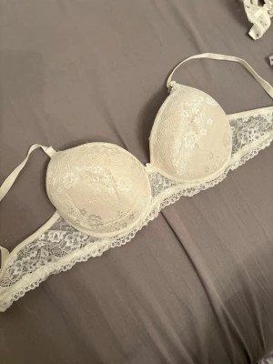Vit spets-bh med vadderade kupor - Snygg vit bh med vadderade kupor och vacker spetsdetalj runt om. Justerbara axelband och bygel för extra stöd. Perfekt för dig som gillar en feminin och romantisk stil. Materialet är en mix av spets och slät textil.