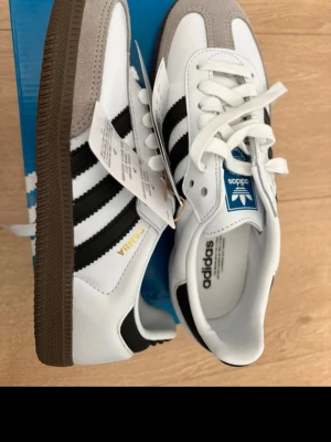 Adidas Samba OG vita och svarta sneakers storlek 36 1/2  - Klassiska Adidas Samba OG sneakers i vitt läder med svarta ränder och grå mockadetaljer på tån. Köptes nyligen men det passade ej. Priset går att diskutera. 