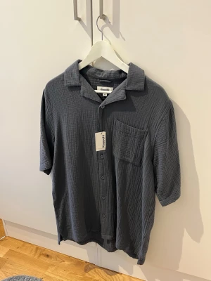 Grå/Blå KappAhl Skjorta Oanvänd - Säljer en mörkgrå skjorta från Kappahl med korta ärmar och avslappnad passform. Skjortan har en bröstficka, knappar framtill och är gjord i ett mjukt, strukturerat bomullstyg. Perfekt för en chill och stilren look.