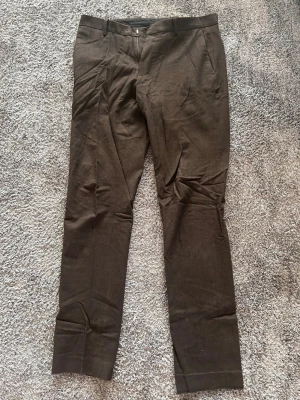 Bruna kostymbyxor/ chinos med rak passform - Snygga bruna kostymbyxor med rak passform och klassiska pressveck. Byxorna har bälteshällor, framfickor och stängs med dragkedja och knapp. Perfekta för en stilren och modern look. Materialet känns mjukt och har en lätt struktur.