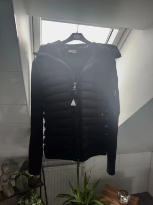 Svart dunjacka med huva från Moncler - Snygg svart dunjacka från Moncler med quiltad front, stickade ärmar och huva. Jackan har dragkedja framtill och en liten Moncler-logga på ärmen. Perfekt för kalla vinterdagar och riktigt stilren look. Observera att De är en 1:1 jacka som ny har används 2 gånger 