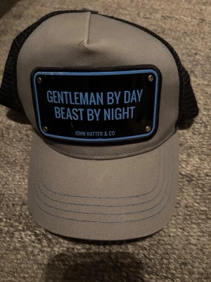 Grå truckerkeps John Hatter & Co - Snygg grå truckerkeps från John Hatter & Co med svart mesh baktill och böjd skärm. Framtill sitter en svart skylt med blå text: 'Gentleman by day, beast by night'. Perfekt statement piece för dig som vill sticka ut.