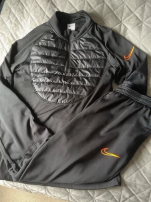 Svart Nike tröja med quiltad front - Snygg svart tröja från Nike med quiltad, vadderad frontpanel och halv dragkedja. Orange Nike-logga på ärmen ger en sportig vibe. Tillverkad i mjukt material, perfekt för lager-på-lager och streetwear. Passar dig som gillar sportig och modern stil. Pris kan ej diskuteras, helt ny och oanvänd. Ställ gärna frågor ifall om ni tänker köpa