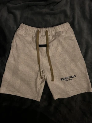 Essentials  shorts grå - Grå shorts från Essentials Fear of God med elastisk midja och långa olivgröna snören framtill. Tryckt logga på vänster ben. Sköna och stilrena shorts i mjukt bomullsmaterial, perfekta för avslappnade dagar.