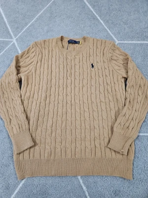 Beige kabelstickad tröja Polo Ralph Lauren - Kabelstickad tröja i beige från Polo Ralph Lauren med klassisk marinblå logga broderad på bröstet. Tröjan har rund halsringning, ribbade muddar och nederkant. Perfekt för dig som gillar tidlös och stilren design.