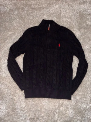 Svart kabelstickad tröja Polo Ralph Lauren - Kabelstickad svart tröja från Polo Ralph Lauren med halv dragkedja och hög krage. Tröjan har det klassiska röda Polo-logot broderat på bröstet och ribbade muddar vid ärmslut och nederkant. Perfekt för en stilren och avslappnad look.
