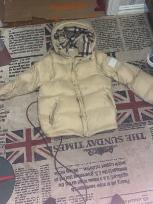 Beige pufferjacka från Burberry - Snygg beige pufferjacka från Burberry med klassiskt rutigt foder i huvan och tydlig Burberry London England-patch på ärmen. Jackan har dragkedja, tryckknappar och två stora fickor framtill. Perfekt för kalla vinterdagar och riktigt trendig look. Storlek L men är som en M passar mig bra är 183 och 65 kg
