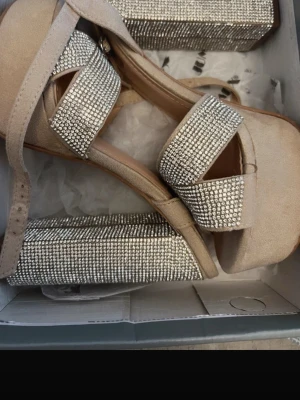 Beige platåsandal med strassdetaljer - Säljer ett par beigea platåsandaler med breda band täckta av glittrande strass. Sandalerna har en hög platåsula och justerbart ankelband. Perfekta för dig som vill sticka ut med extra bling. Materialet är mockaimitation och syntet.