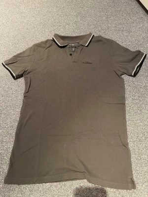 Grå pikétröja från H&M - Snygg grå pikétröja från H&M med klassisk krage och knäppning framtill. Tröjan har svarta och vita ränder på kragen och ärmarna för en sportig look. Perfekt till jeans eller shorts.