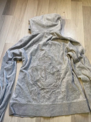Grå hoodie från Juicy Couture - Står storlek XL men känns som s. Säljer en grå hoodie från Juicy Couture med dragkedja framtill och huva. Baksidan har ett stort broderat Juicy Couture-emblem med krona och JC-logga. Hoodien har ribbade muddar och fickor framtill. Perfekt för en chill och trendig look. 
