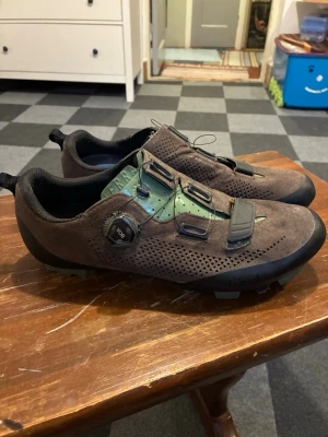 Bruna cykelskor från Fizik - Säljer ett par bruna cykelskor från Fizik med perforerat mockaläder, gröna detaljer och BOA-snörning för smidig justering. Skorna har robust sula och förstärkta partier vid tår och häl, perfekta för MTB eller gravel.