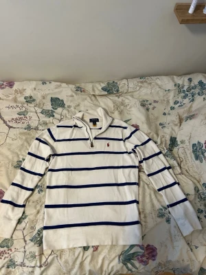 Vit stickad tröja med blå ränder Polo - Vit stickad tröja från Polo Ralph Lauren med marinblå horisontella ränder och liten röd logga på bröstet. Tröjan har halv dragkedja vid halsen, lång ärm och ribbade muddar. Perfekt för en avslappnad och stilren look. 