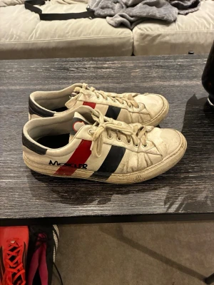 Moncler vita sneakers med röd och blå detalj - Snygga vita sneakers från Moncler med klassiska röd och marinblå ränder på sidan samt svart hälparti. Skorna har lågt skaft, vita snören och en stilren design som passar till det mesta. Tillverkade i läder med textildetaljer och tydlig Moncler-logga på sidan. Riktigt risigt skick på dessa men hålet under är lätt fixat genom att gå till en skomakare eller bara sätta nån tejpbit. Äkta skor