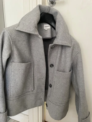 Grå cardiganjacka från Kappahl - Stilren grå cardiganjacka från Kappahl med stora framfickor och knappar framtill. Jackan har en bred krage och är gjord i ett mjukt, filtliknande material. Perfekt för lager på lager och enkel att matcha med olika outfits 