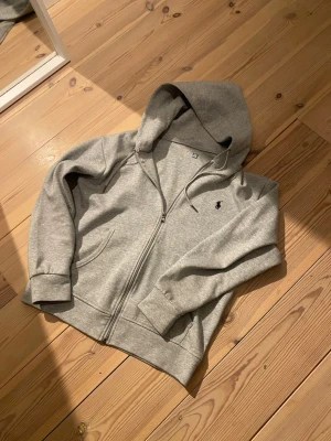 Grå hoodie från Polo Ralph Lauren - Snygg grå hoodie från Polo Ralph Lauren med dragkedja framtill och klassisk huva. Tillverkad i mjuk bomullsblandning och har ribbade muddar vid ärmar och nederkant. Liten broderad logga på bröstet och två fickor framtill.