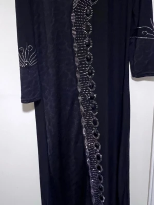 Svart långklänning med pärldetaljer - Elegant svart långklänning med dekorativa pärlor i ett vågigt mönster framtill och på ärmarna. Klänningen har trekvartsärm och rak passform, vilket ger en stilren och sofistikerad look. Perfekt för dig som gillar unika detaljer.