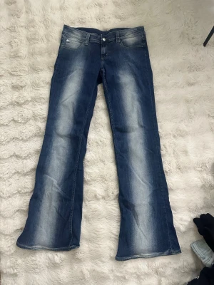 Blå utsvängda jeans som har ljusa partier fram och bak. - Snygga blå bootcut jeans med ljusa slitningar framtill och klassisk femficksdesign. Jeansen har låg midja och stängs med knapp och dragkedja.