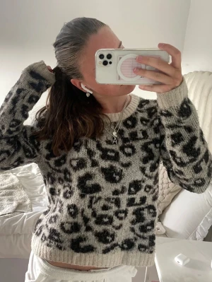 Beige leopardmönstrad stickad tröja - En beige stickad tröja med svart leopardmönster. Tröjan har rund halsringning och långa ärmar med ribbade muddar. Materialet ser ut att vara mjukt och lite fluffigt, perfekt för att hålla sig varm. Passar dig som gillar trendiga mönster och comfy stil.