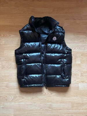 Svart dunväst från Moncler - Snygg svart dunväst från Moncler med glansig finish och klassisk logotyp på bröstet. Västen har hög krage, två fickor med dragkedja och quiltad design. Perfekt för lager-på-lager och håller dig varm under kyliga dagar.