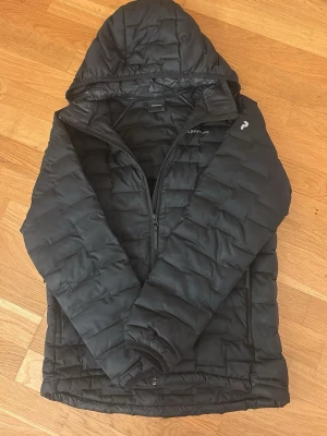 Svart dunjacka från Peak Performance - Snygg svart dunjacka från Peak Performance med huva och dragkedja framtill. Jackan har två sidofickor med dragkedja. I storlek S/170 och har en lagnings lapp på baksidan som suttit där ett tag byts den ut så kommer den knappt synas! Syns inte på långt avstånd.