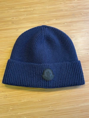 Mörkblå Moncler mössa i ull - Snygg mörkblå ribbstickad mössa från Moncler med uppvikt kant och Moncler-logga framtill. Tillverkad i mjuk ull som håller dig varm och ger en clean look. Perfekt accessoar för kalla dagar och enkel att matcha med olika outfits.