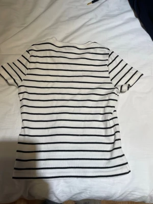 Vit t-shirt med svarta ränder - Säljer en vit t-shirt med tunna svarta horisontella ränder. T-shirten har rund halsringning och korta ärmar. Materialet är ribbat och stretchigt vilket ger en skön passform. Perfekt till jeans eller kjol för en enkel och stilren look.