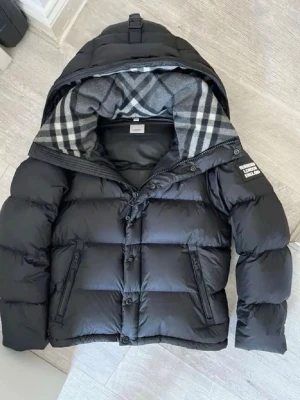 Svart pufferjacka från Burberry - Snygg svart pufferjacka från Burberry med stor huva och klassiskt rutigt foder. Jackan har dragkedja och knappar framtill, två sidofickor och en patch med Burberry-logga på ärmen. Perfekt för kalla vinterdagar och riktigt trendig look.