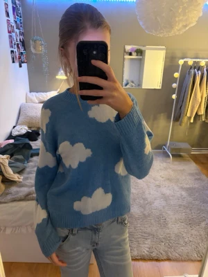Blå stickad tröja med molnmönster - Supermysig blå stickad tröja med vita moln över hela. Tröjan har rund halsringning, långa ärmar och ribbstickade muddar. Perfekt för dig som gillar unika plagg. Materialet känns mjukt och fluffigt, perfekt för svala dagar.