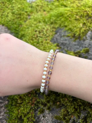 Två pärlarmband i vitt och lila - Säljer två stretchiga armband med små pärlor. Ett armband har vita pärlor och det andra har genomskinliga lila pärlor. Perfekta att bära tillsammans eller var för sig för en enkel men snygg look.