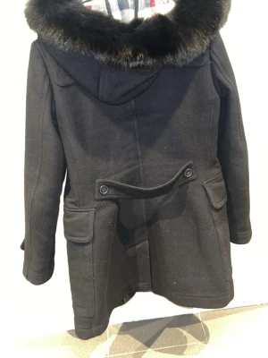 Svart Burberry Brit kappa med päls - Stilren svart kappa från Burberry Brit med huva och fuskpälsdetalj runt kragen. Kappan har klassiskt rutigt foder, fickor med lock och snygga knapprader bak. Perfekt för kyliga dagar när du vill vara både varm och trendig. Pris går att diskutera.