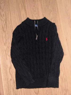 Svart kabelstickad tröja Polo Ralph Lauren - Kabelstickad svart tröja från Polo Ralph Lauren med halv dragkedja och röd broderad logga på bröstet. Tröjan har ribbade muddar och hög krage, perfekt för lager på lager. Klassisk och stilren design i mjukt material.