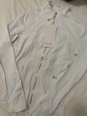 Vit träningsjacka från Lululemon st 4 - Snygg vit träningsjacka från Lululemon med guldiga dragkedjor och figurnära passform. Jackan har långa ärmar, hög krage och är tillverkad i ett stretchigt syntetmaterial som passar perfekt för träning. Stilsäker och sportig design med diskreta sömdetaljer.
